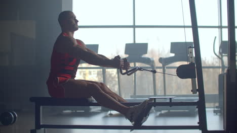 Young-man-doing-workouts-on-a-back-with-power-exercise-machine-in-a-gym-club.-At-athletic-man-doing-workouts-on-a-back-with-power-exercise-machine-in-a-gym-on-the-background-of-large-Windows.